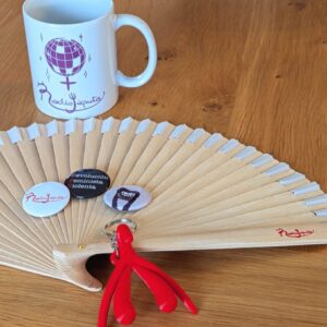 Un abanico de madera con el logo en rojo de Radiojaputa en el lateral. Una taza con el texto "feminista reina de la pista" y una imagen de una bola de discoteca. Un llavero rojo con forma de clítoris y labios. 3 chapas.