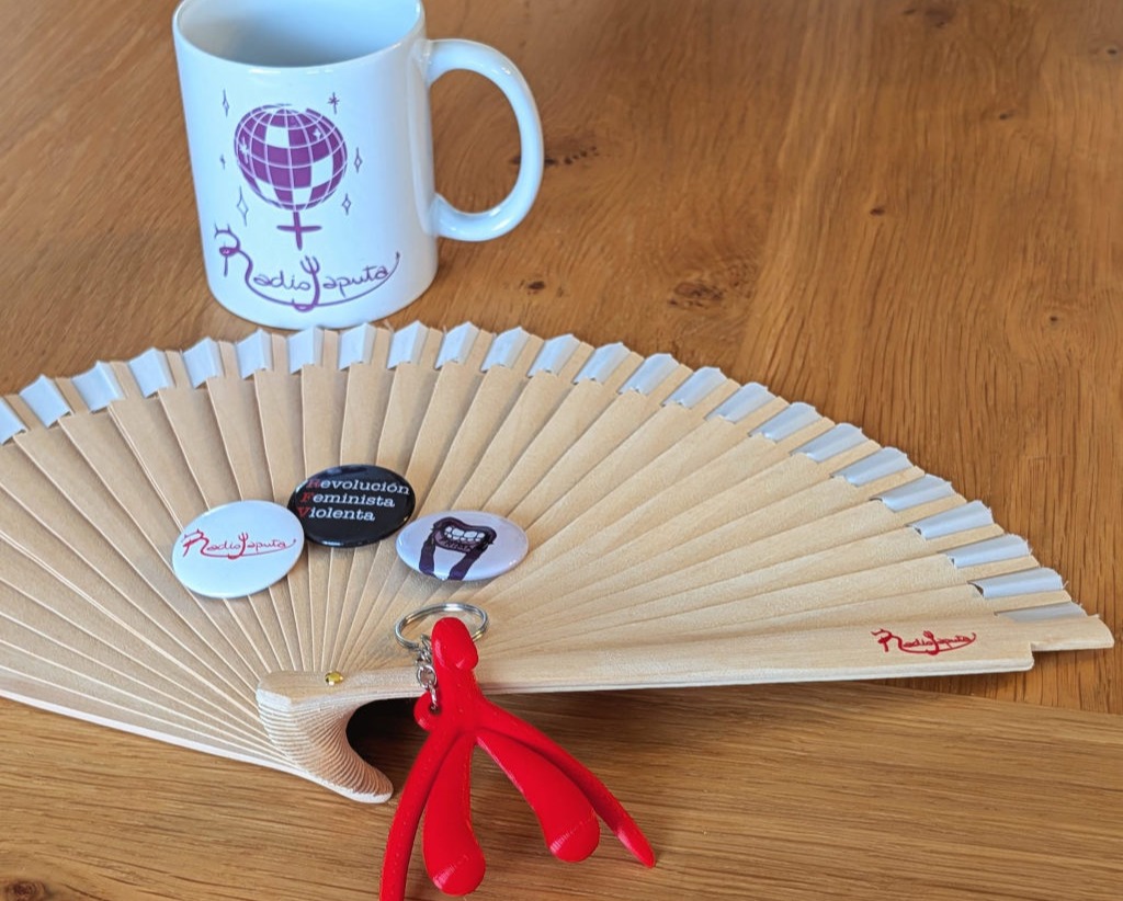 Un abanico de madera con el logo en rojo de Radiojaputa en el lateral. Una taza con el texto "feminista reina de la pista" y una imagen de una bola de discoteca. Un llavero rojo con forma de clítoris y labios. 3 chapas.